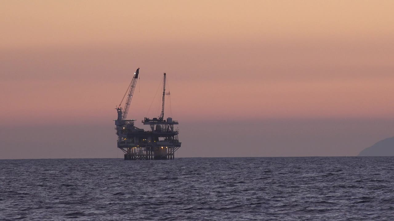 Offshore oil platform near Huntington Beach sits left of center on dark ocean water under a pink sunset sky, with no visible people.