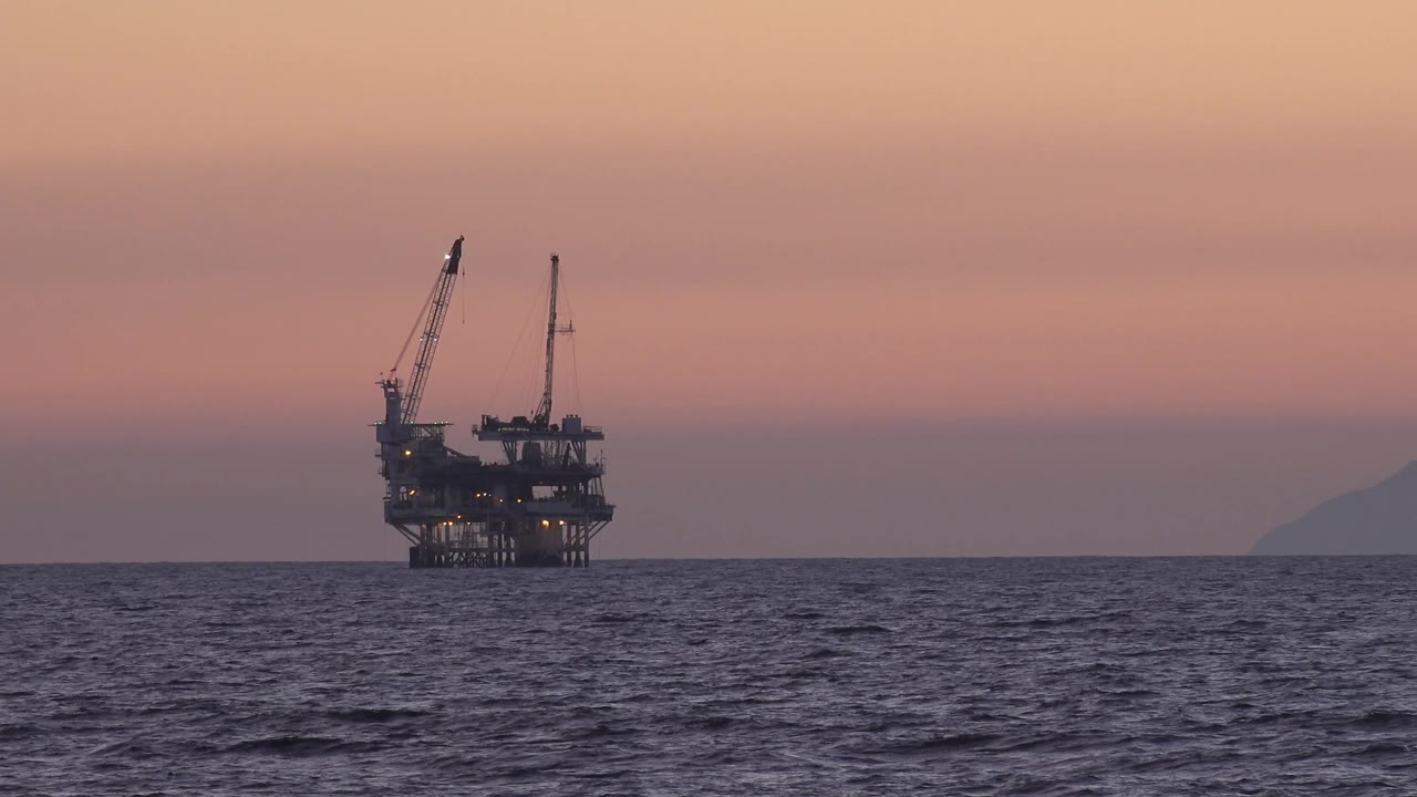 A distant offshore platform with crane booms stands against a muted sunset sky near Huntington Beach, with the coastline faintly visible at the right edge and no visible people.