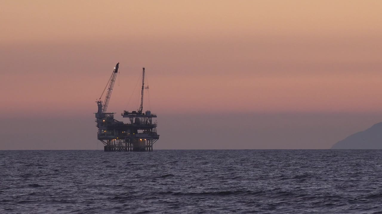 Offshore platform with two cranes sits left of center over dark ocean water in Huntington Beach, California, with pink dusk sky, a distant landform on the right, and no visible people.