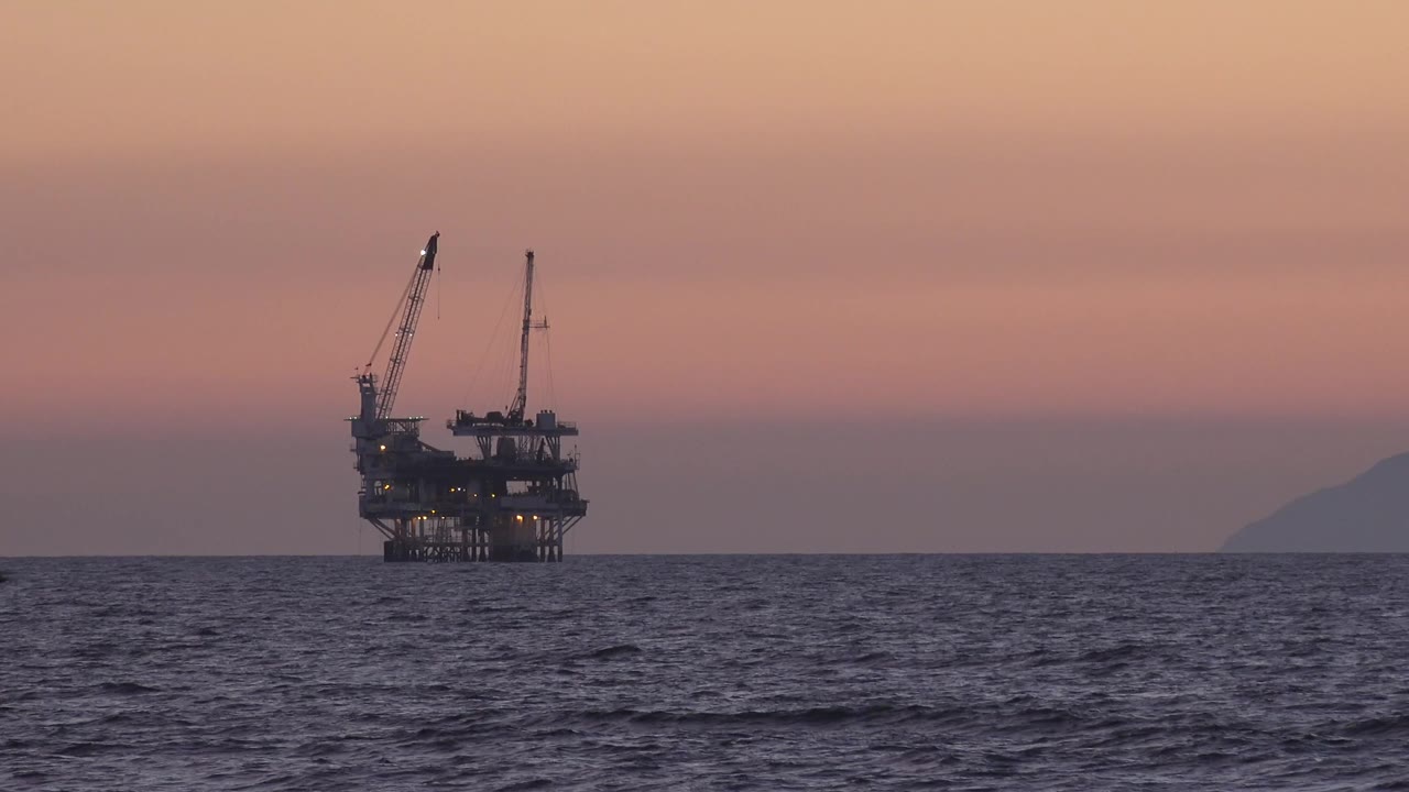 The platform remains near the left side of the frame in Huntington Beach, California, with rippled ocean foreground, muted pink sky, a right-horizon landform, and no visible people.