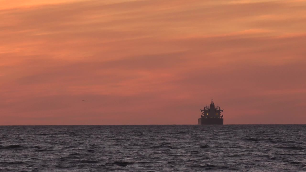A distant boat sits near the Huntington Beach ocean horizon under an orange-pink sky, with dark rippled water below and no visible people.