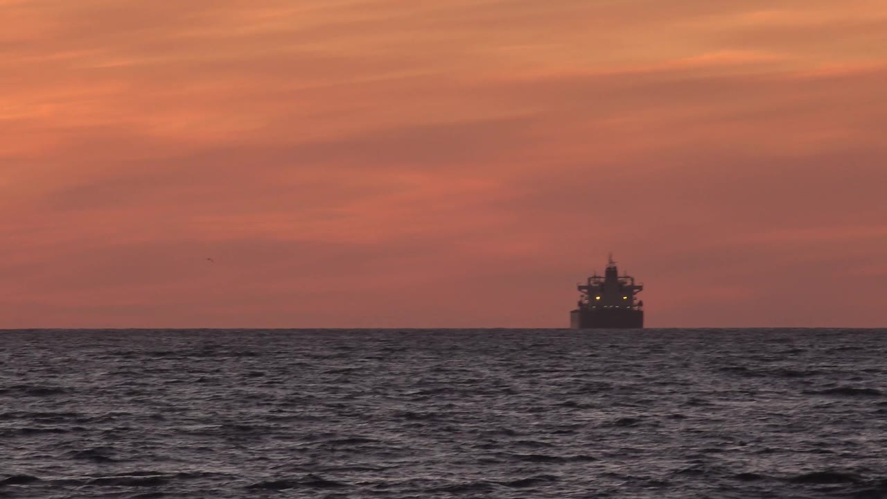 A dark vessel sits near the right side of the Huntington Beach sea horizon, with gray waves in front and warm cloud bands above, and no visible people.