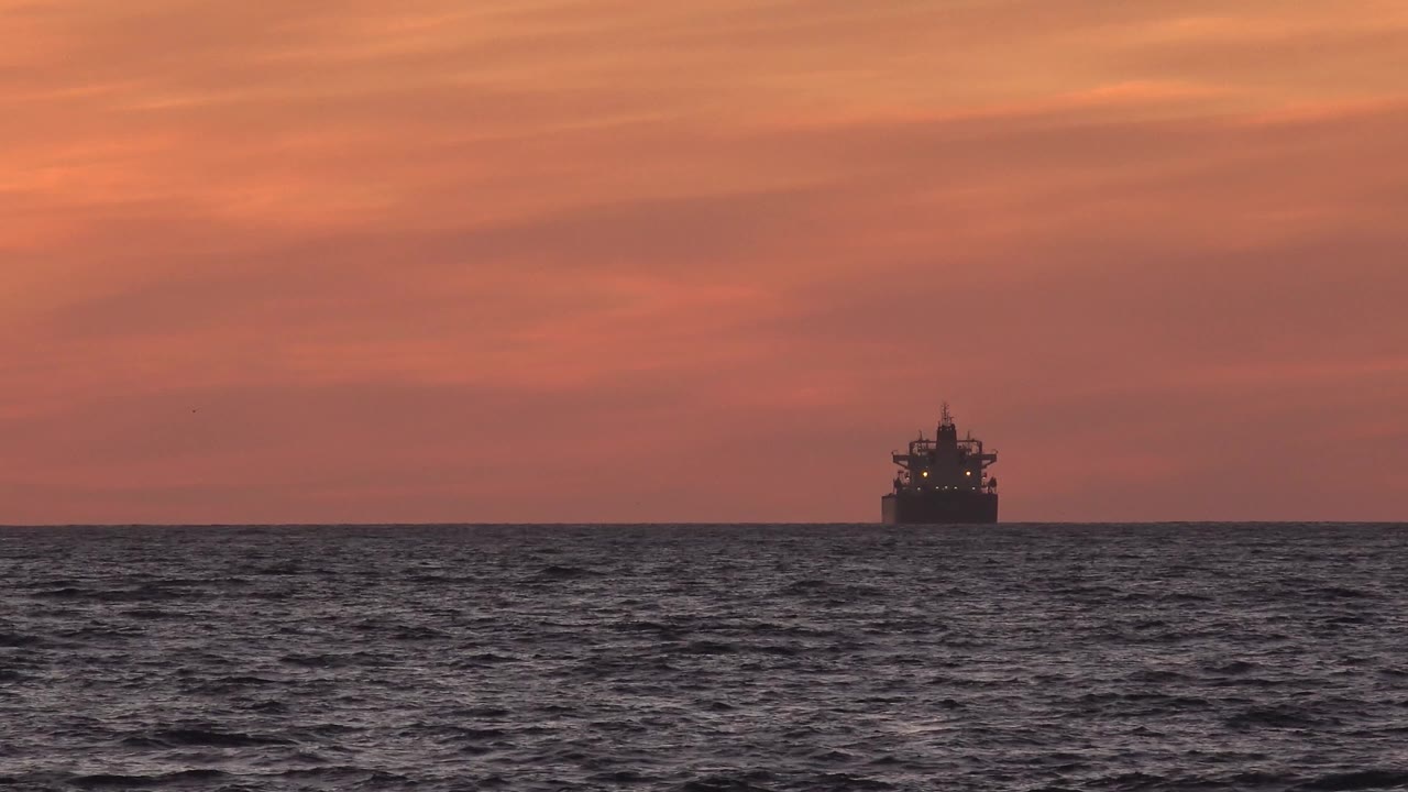 A distant tanker silhouette sits near the center of the Huntington Beach ocean horizon under an orange sunset sky, with dark rippled water in front and no visible people.