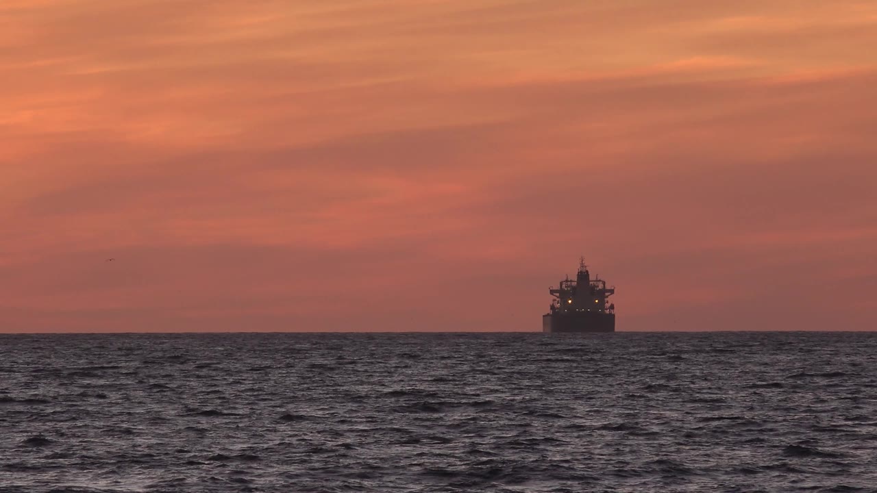 A small dark tanker appears toward the right side of the Huntington Beach horizon, set against a red-orange sky above choppy ocean water with no visible people.