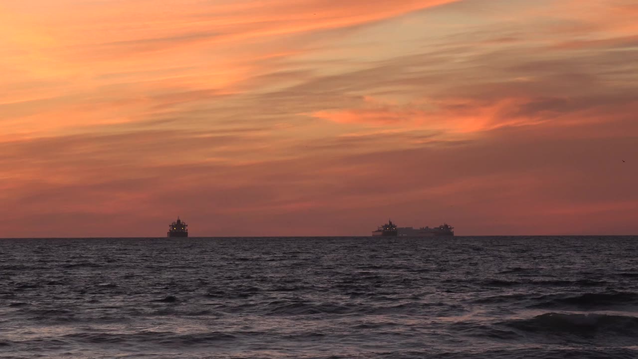 Orange sunset over ocean waves with distant ship silhouettes.