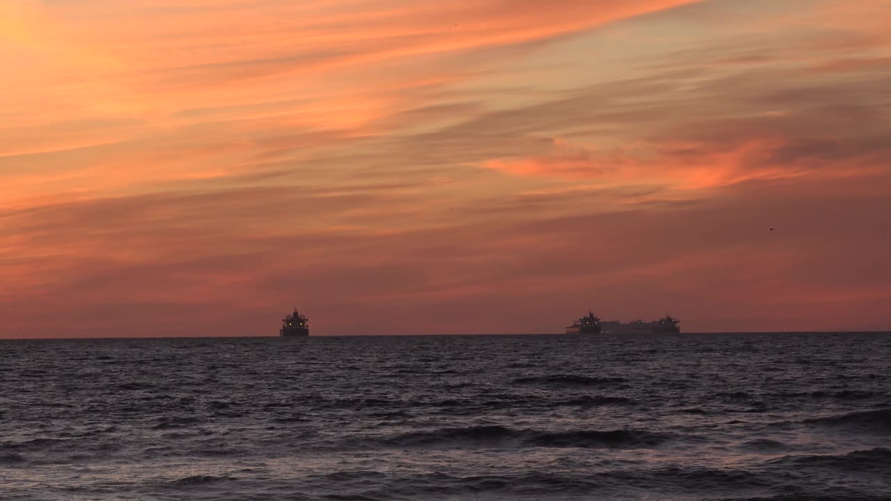 Huntington Beach frame with a darker wave line crossing the foreground, distant ship silhouettes near the horizon, orange cloud layers, and no visible people.