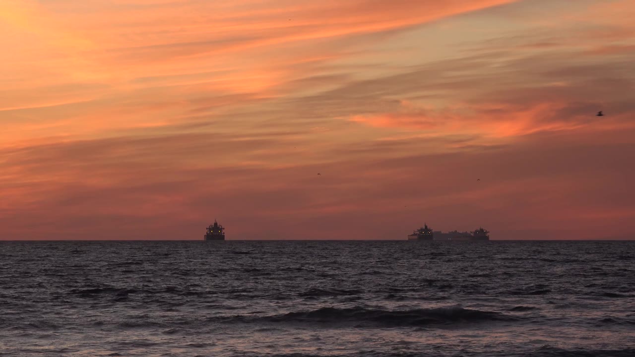Huntington Beach sunset frame with a darker band of surf in the foreground, broad orange clouds above, and distant grey horizon objects, with no visible people.