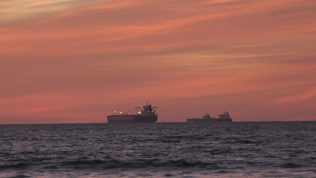 At 0:01, the two cargo ships remain separated along the Huntington Beach horizon as small waves darken the lower frame, with no visible people.