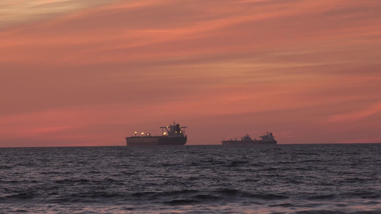 At 0:02, two silhouetted cargo ships are visible beyond choppy water off Huntington Beach, California, under a red-orange sky with no visible people.