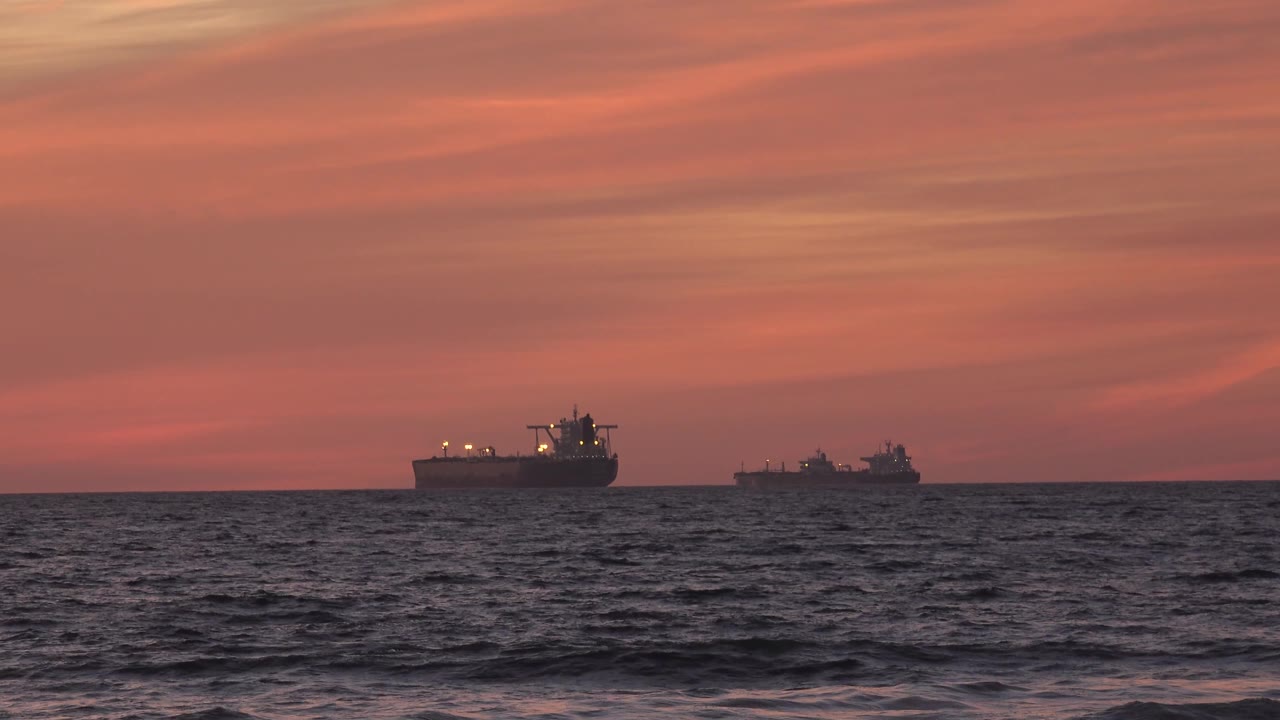 At 0:03, the larger cargo ship appears left of a smaller distant vessel on the Huntington Beach horizon, above textured waves and with no visible people.