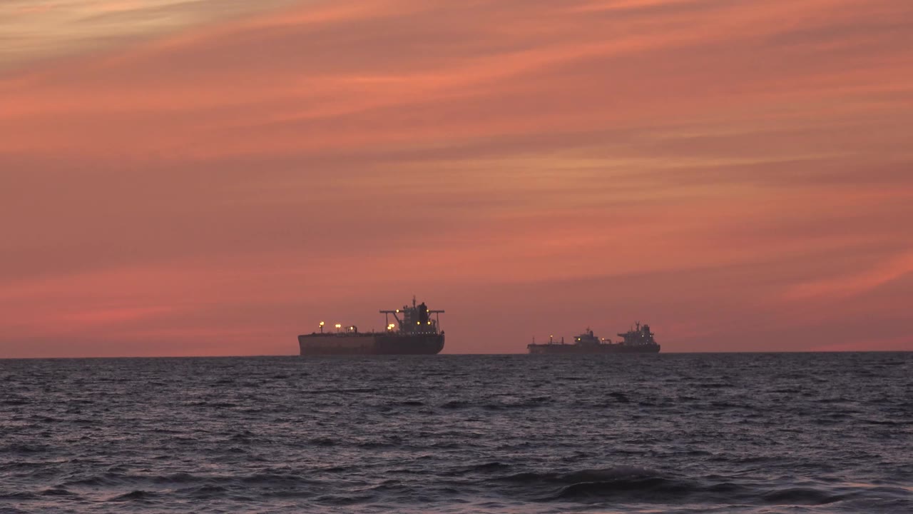 The ships remain near the horizon off Huntington Beach as small waves rise across the foreground beneath streaked sunset clouds, with no visible people.