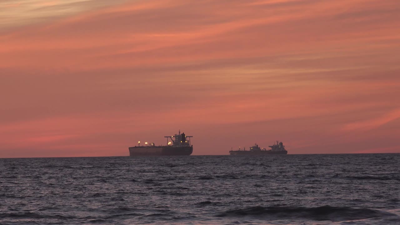 Two vessels are visible beyond the dark sea at Huntington Beach, with warm red-orange cloud bands filling most of the frame and no visible people.