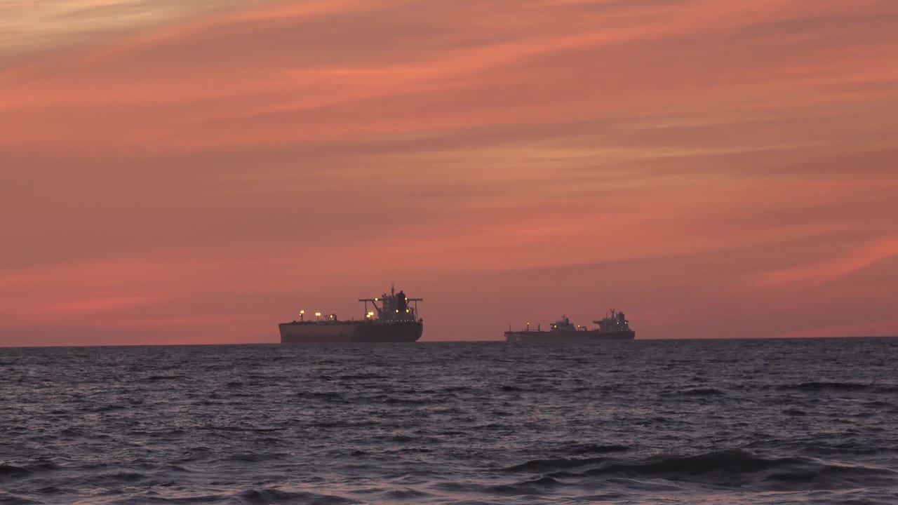 The two ships hold position off Huntington Beach while low foreground waves darken the bottom of the frame under a muted orange sunset sky, with no visible people.