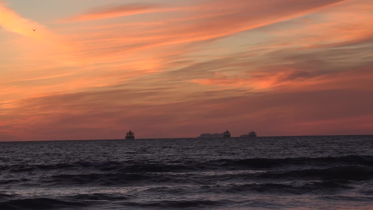 Rolling surf darkens the foreground at Huntington Beach, California, while three small ships sit under orange and pink cloud bands with no visible people.