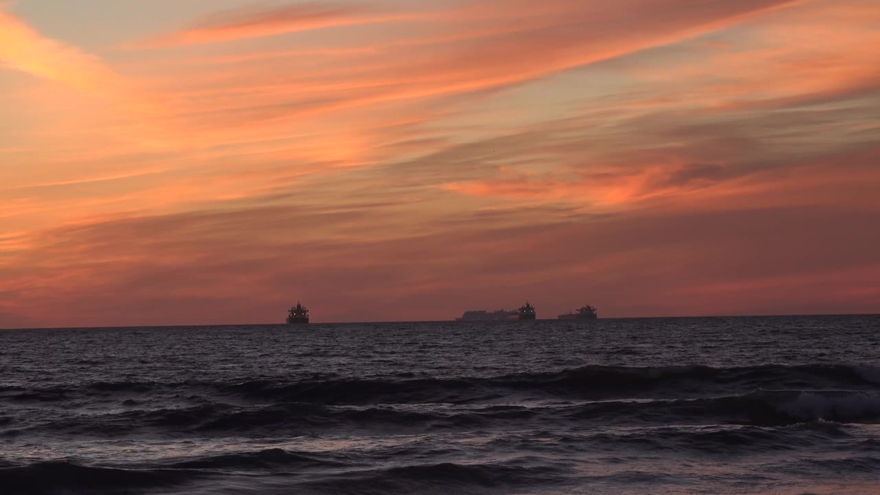 Dark Huntington Beach waves run across the foreground, with silhouetted ships spread along the horizon beneath a warmer orange cloud layer and no visible people.