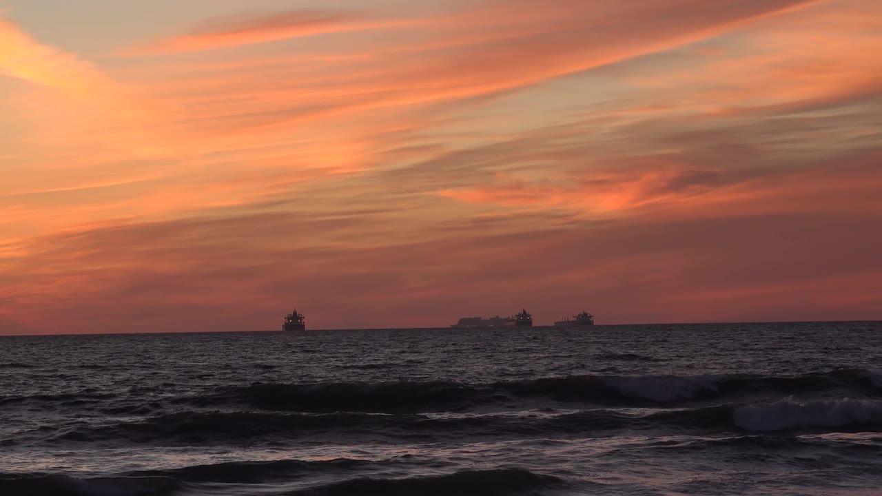 A lower band of surf crosses the Huntington Beach foreground as three distant ships remain on the horizon below pink-orange clouds, with no visible people.