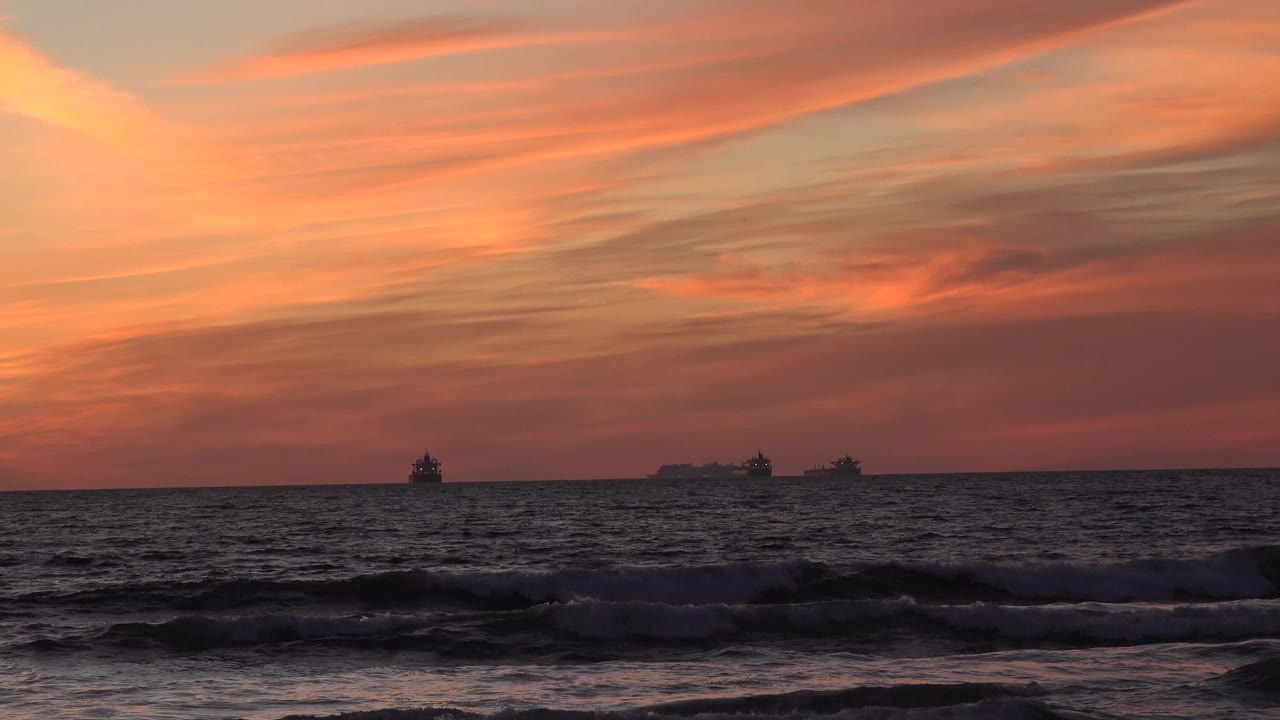 Pink and orange sunset clouds over Huntington Beach ocean waves, with small distant boats on the horizon and no visible people.