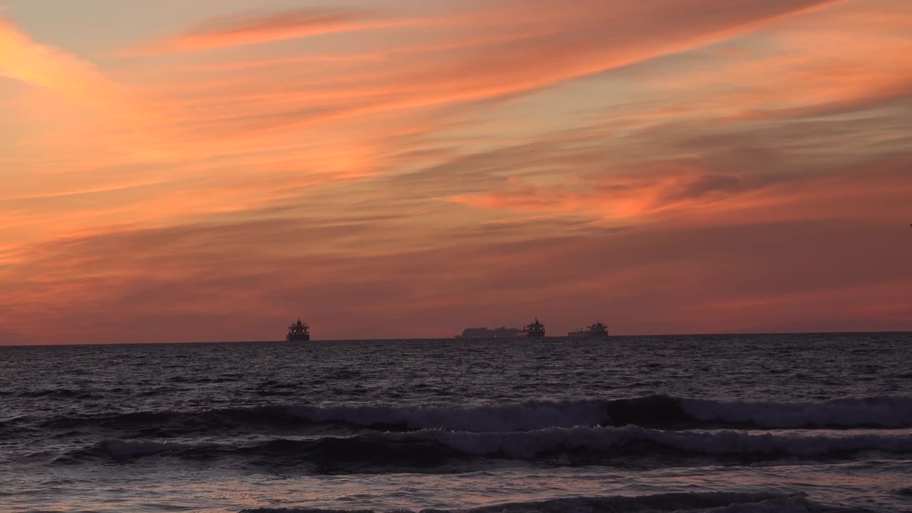 Orange and rose dusk sky stretches over Huntington Beach ocean waves, with several small vessels silhouetted far offshore and no visible people.