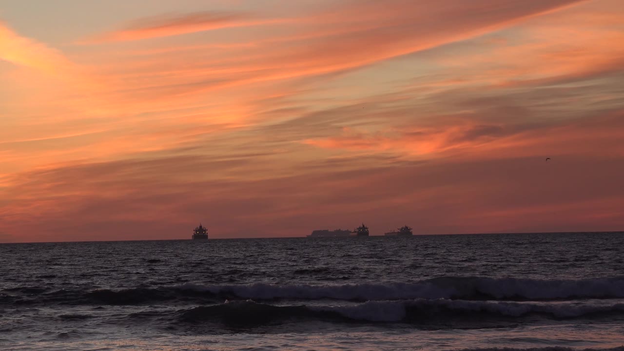 A wider-looking moment of sunset sky over the Huntington Beach sea shows choppy water, low foreground waves, distant boat shapes, and no visible people.
