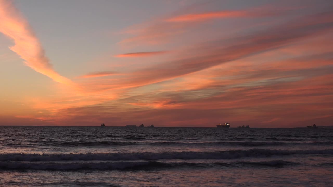 Huntington Beach surf rolls across the foreground while the orange-pink cloud band widens over the horizon, with small distant vessels and no visible people.
