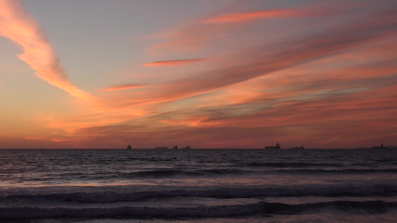 Huntington Beach sunset with dark foreground surf, a straight ocean horizon, distant ship silhouettes, and red-orange clouds across the sky, no visible people.