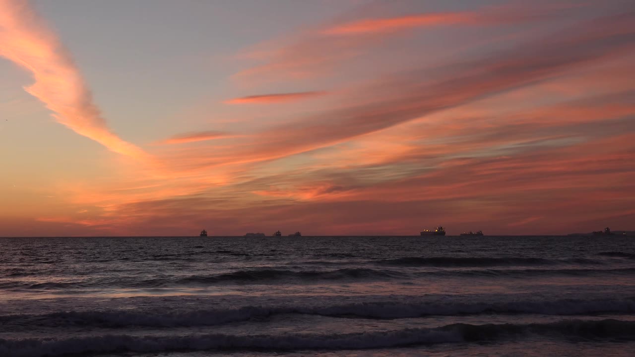 Huntington Beach sunset view with rippled ocean water in the foreground, a thin dark horizon line, small ship silhouettes, and streaked red clouds filling the upper frame, no visible people.
