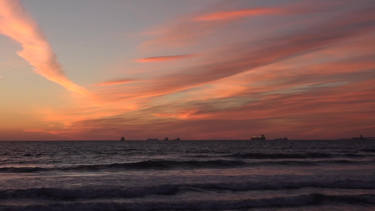 Huntington Beach at sunset with a warm orange glow near the left horizon, dark wave layers in the foreground, and several distant ships under red cloud streaks, no visible people.
