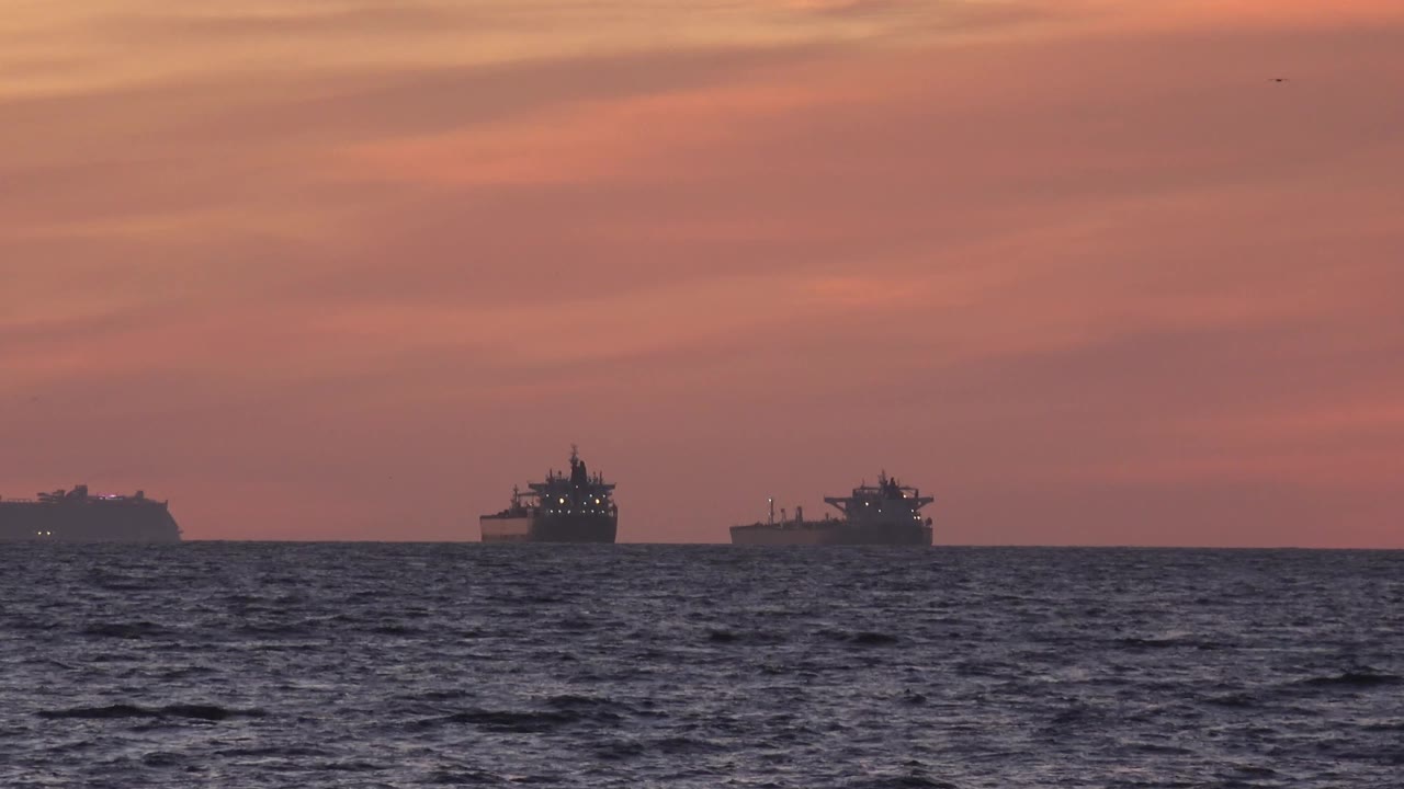 At 0:01, three distant cargo ships line the horizon off Huntington Beach, California, with dark textured water below and soft orange-pink cloud bands above, with no visible people.