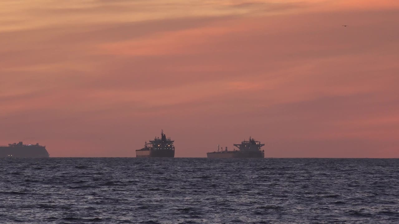 At 0:02, the ships remain spaced across the Huntington Beach, California horizon, with a darker ocean band, muted red sky, and no visible people.
