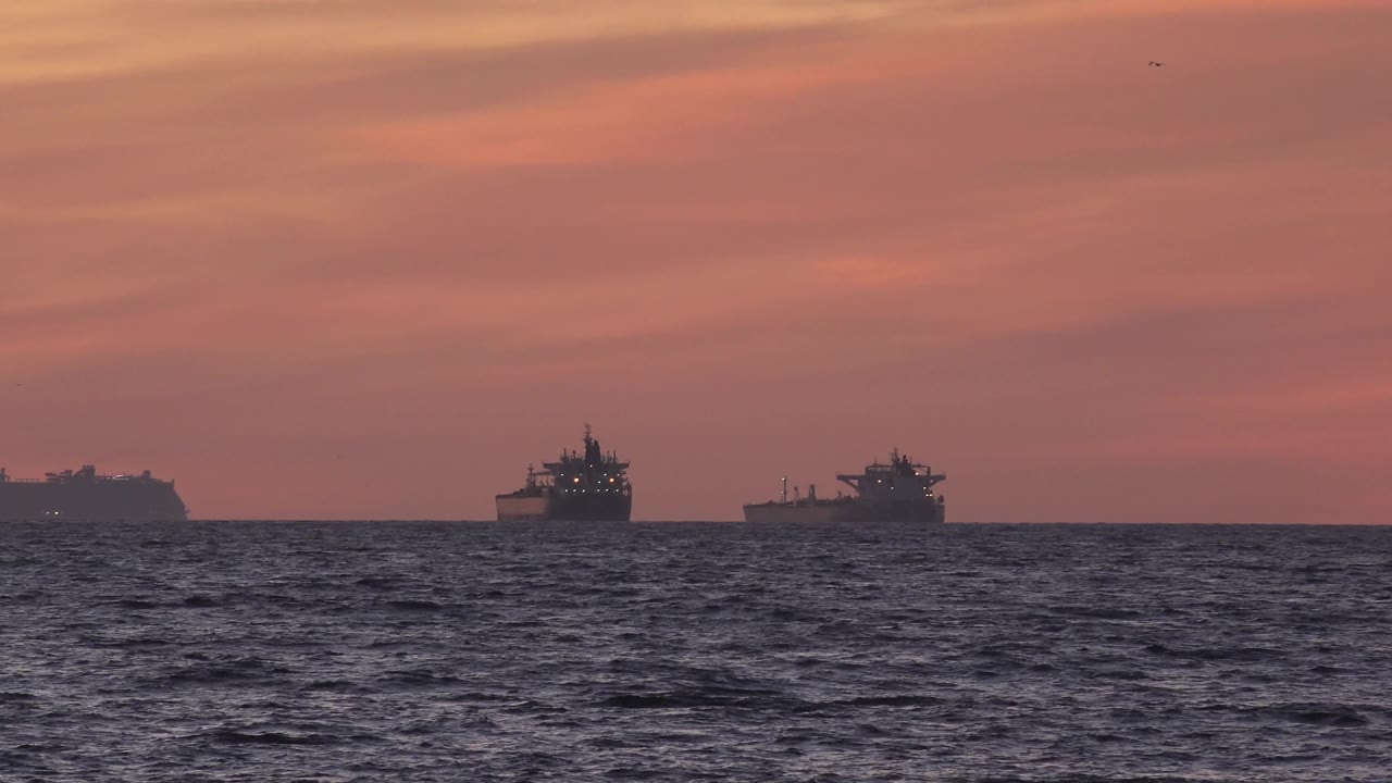 At 0:03, distant cargo ships sit low on the Huntington Beach, California horizon while rolling foreground waves add texture under the pink-orange evening sky, with no visible people.