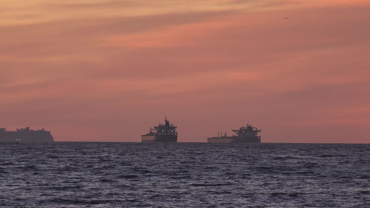 Two dark barges sit on the Huntington Beach ocean horizon under a pink orange sunset sky, with choppy water in the foreground and no visible people.