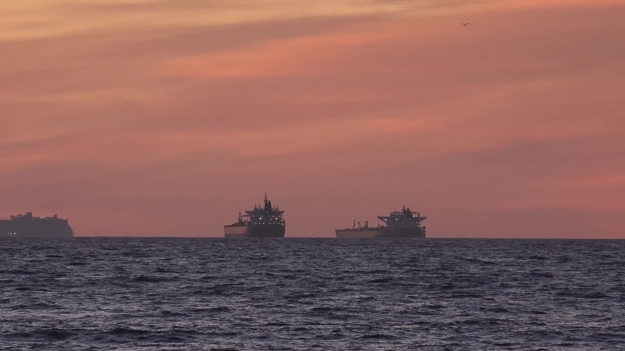 Two distant barges remain visible off Huntington Beach, framed by a broad pink sunset sky and a low strip of textured ocean, with no visible people.