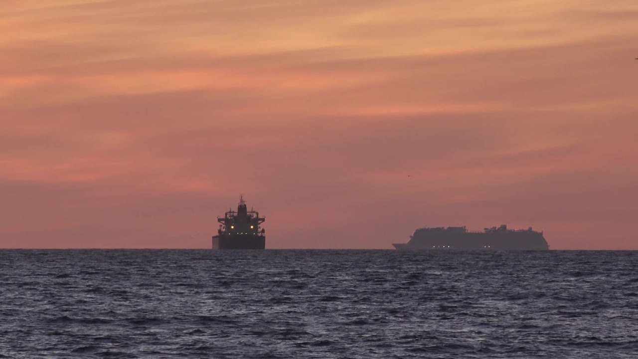 At Huntington Beach, the left ship and larger right vessel remain small dark silhouettes against the red sunset, with choppy ocean in the foreground and no visible people.