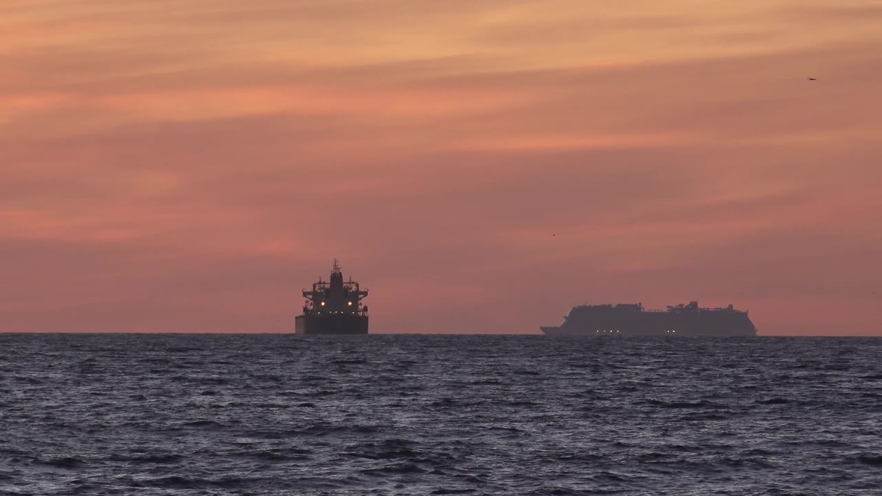 At Huntington Beach, two offshore ship shapes sit low on the horizon below a wide red cloud field, with dark rippled water and no visible people.