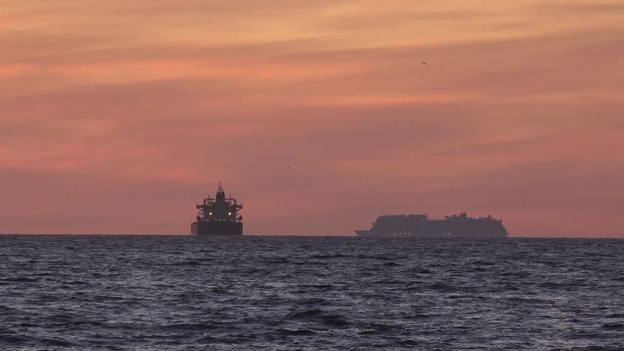 At 0:00, two distant vessel silhouettes sit on the Huntington Beach ocean horizon beneath a pink-orange sunset sky, with textured dark water in front and no visible people.