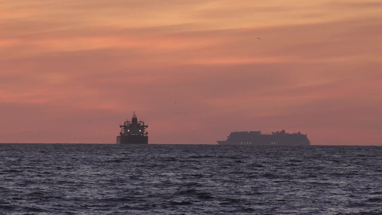 At 0:01, the Huntington Beach horizon holds two dark vessel shapes while small wave patterns move across the foreground water and no visible people appear.
