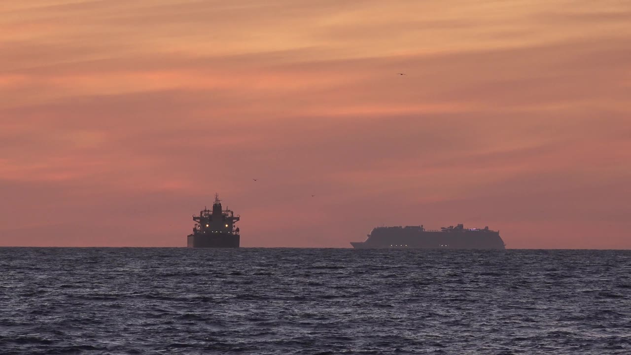 At 0:02, two offshore vessels are silhouetted near the Huntington Beach horizon, with the larger right-hand shape set against warm pink light and no visible people.