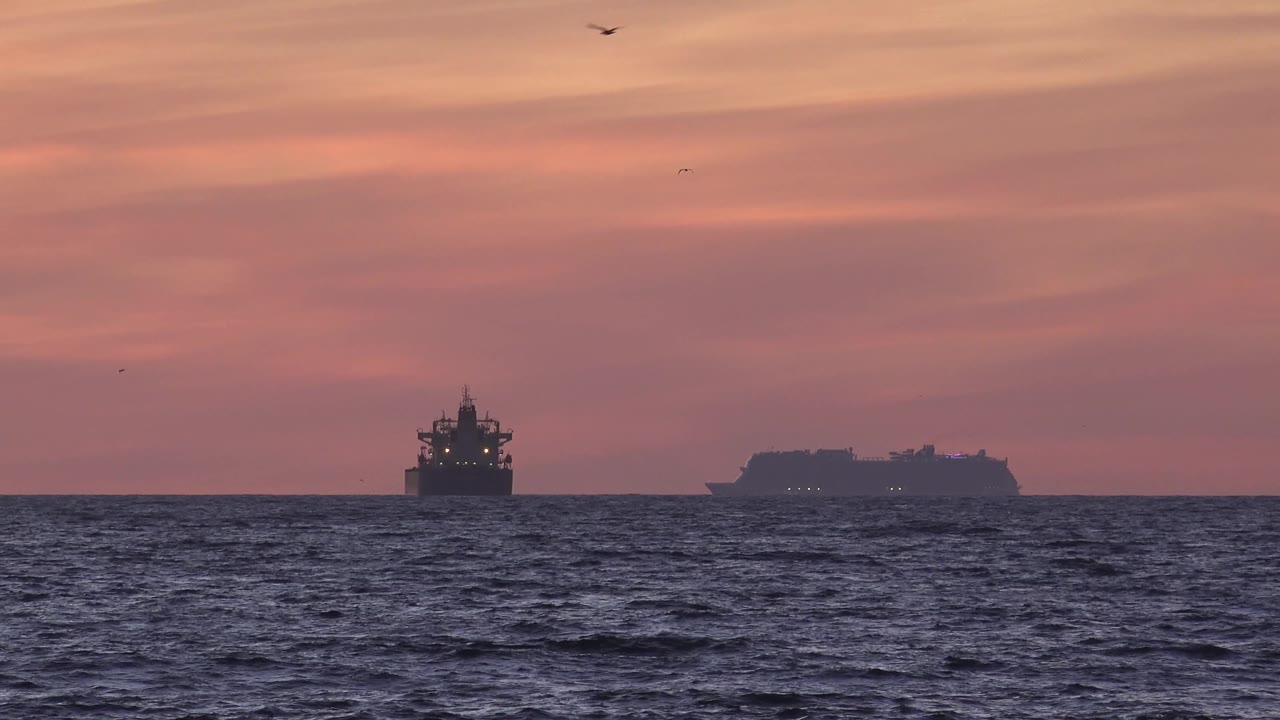 Two offshore vessels are visible from Huntington Beach as dark silhouettes against a muted orange-pink sky, with choppy water in front and no visible people.