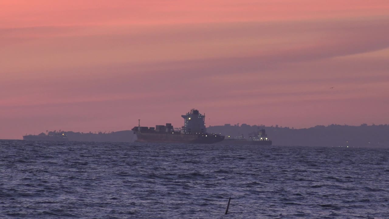 Large offshore ship remains centered near the Huntington Beach horizon, with textured water in front, faint vessel lights nearby, and no visible people.
