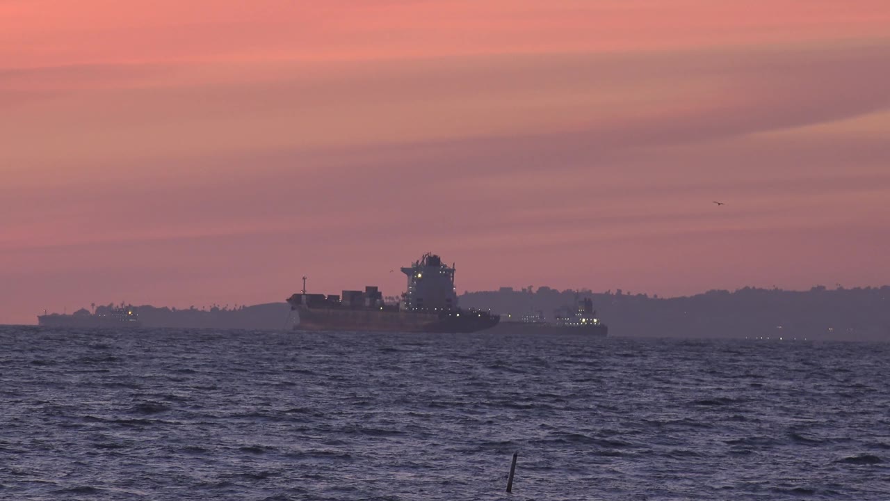 Offshore ship appears below layered pink cloud bands near Huntington Beach, with dark ocean waves in the foreground and no visible people.