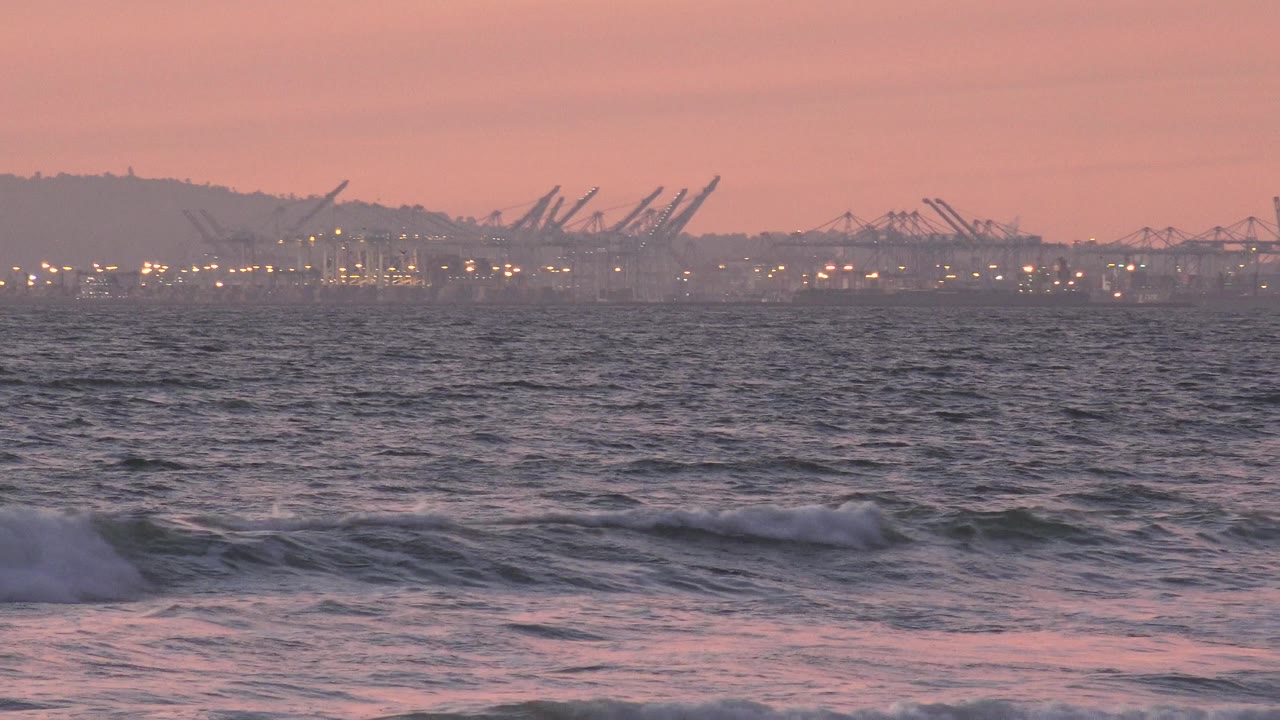 At Huntington Beach, California, small waves cross the foreground while distant port cranes and lights sit under a pink evening sky, with no visible people.