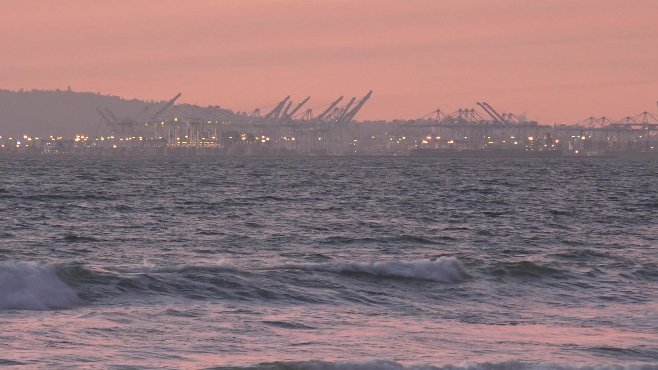 A low wave begins to break across the Huntington Beach foreground as silhouetted cranes and scattered lights remain soft on the far horizon, with no visible people.