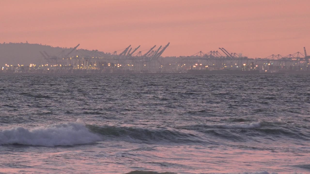 Foam spreads along a small wave in the foreground while the distant Huntington Beach industrial waterfront stays visible through haze, with no visible people.