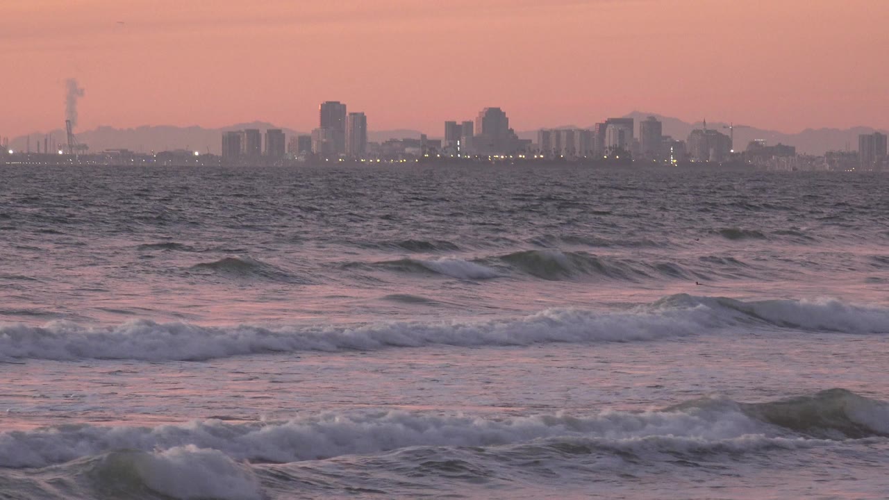 Huntington Beach surf rolls in under pink dusk light with a hazy city skyline on the horizon and no visible people.