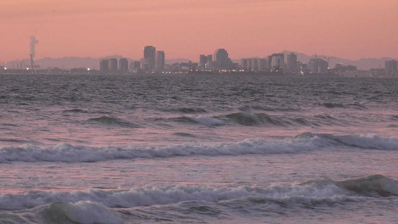 Low surf lines move toward shore beneath a pink sky and skyline.