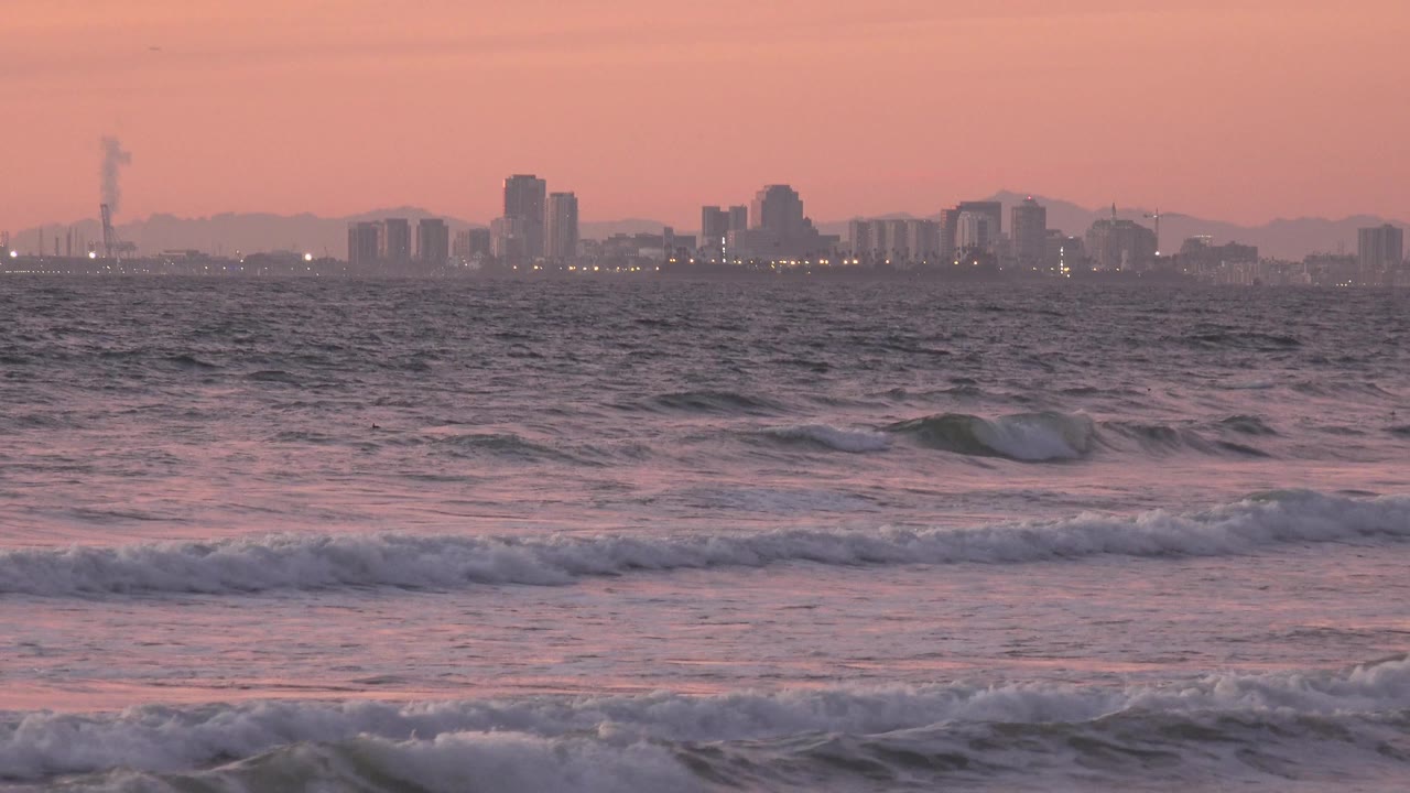 Foam spreads across the near surf as the distant Huntington Beach city skyline sits in soft pink haze with no visible people.