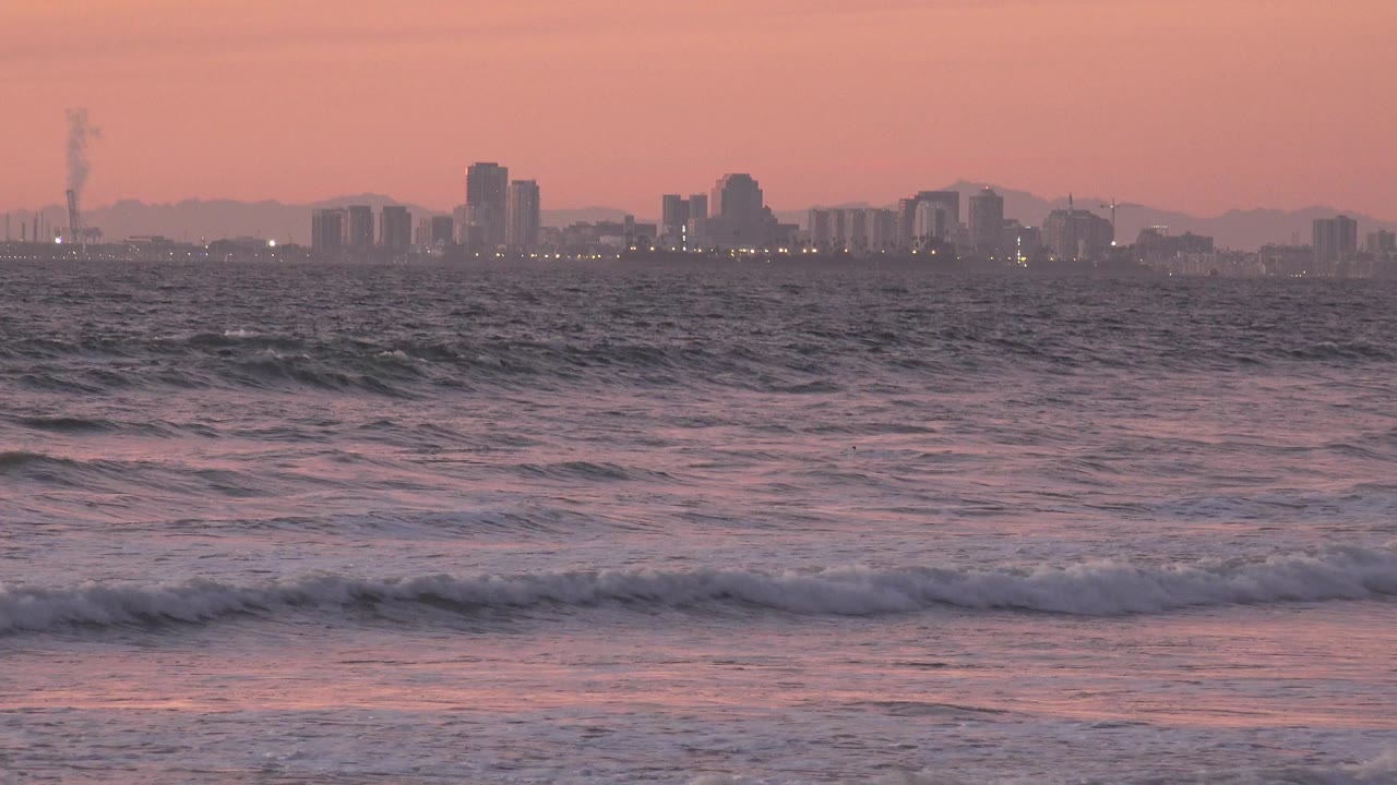 Waves breaking before a distant skyline under a pink sky.