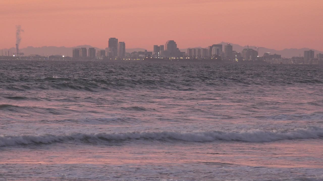 Choppy ocean water and a low foam line fill the foreground while Huntington Beach's distant buildings sit in haze beneath warm sunset color, with no visible people.