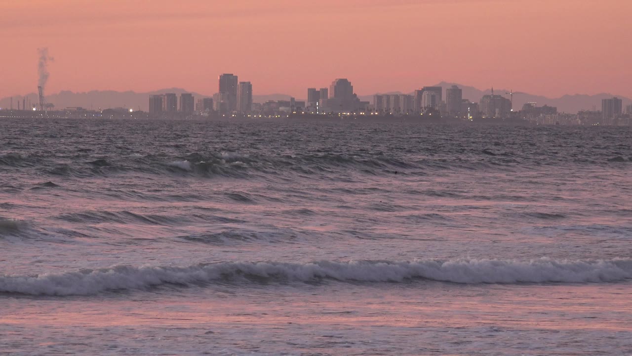 A low wave line rolls toward shore as the hazy Huntington Beach skyline and a narrow smoke plume remain visible across the water, with no visible people.