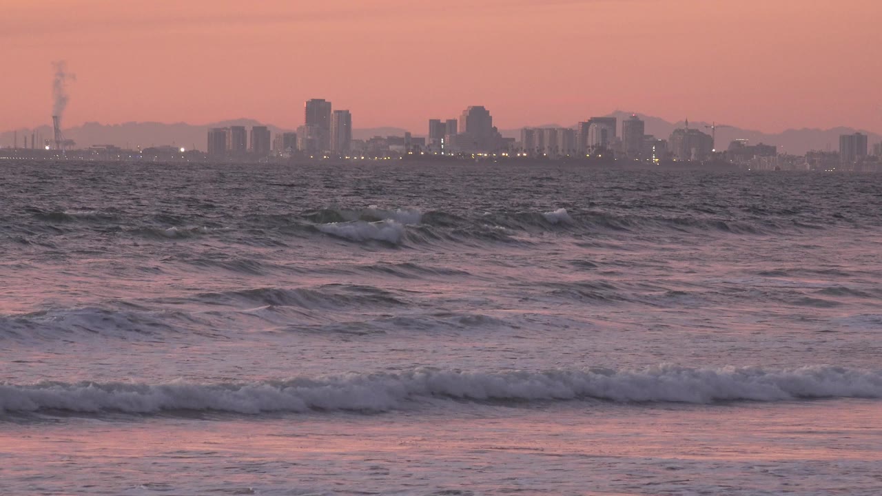 Rough surf rises higher in the foreground while distant high-rise buildings and small shoreline lights sit under a muted pink Huntington Beach sky, with no visible people.